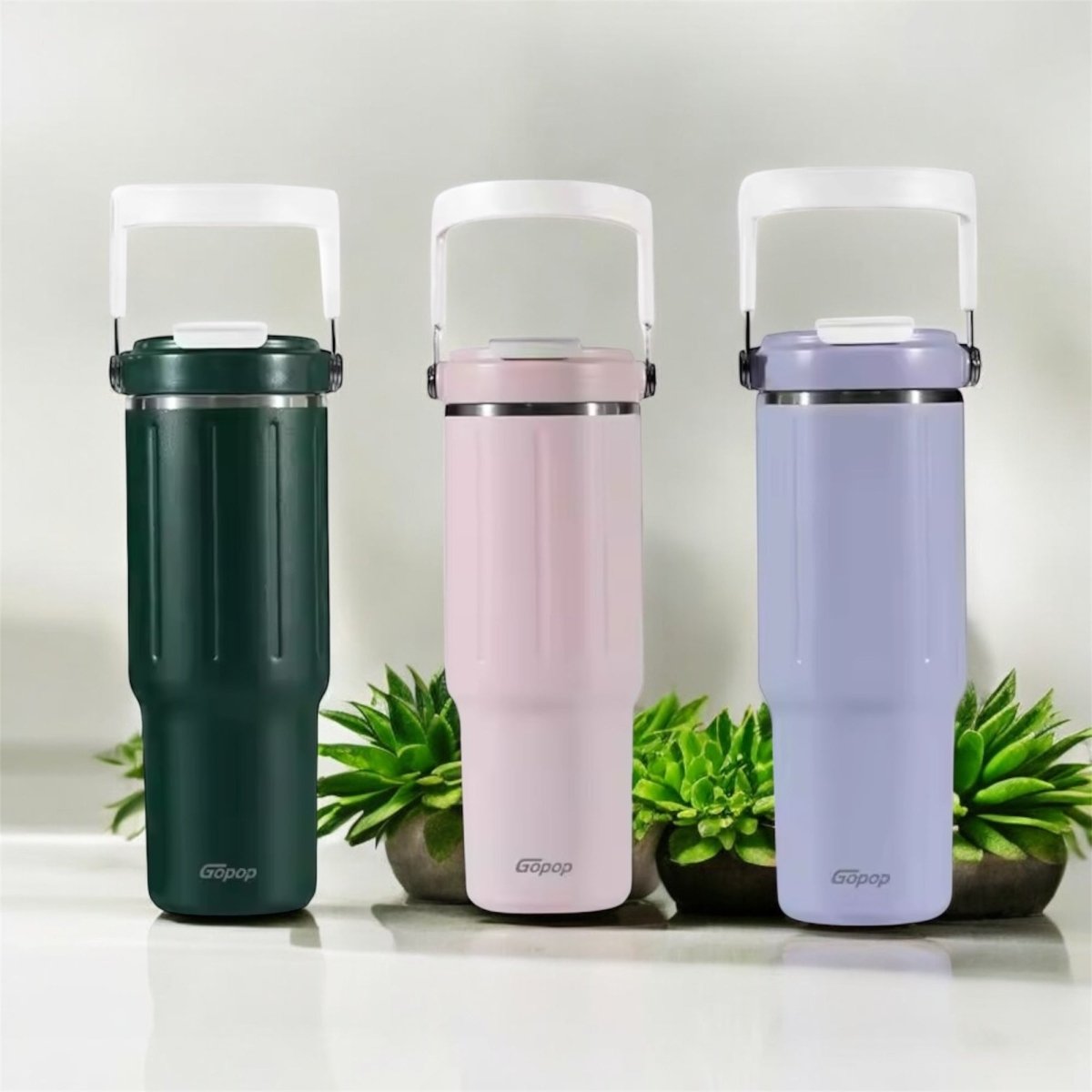 Three insulated travel mugs in green, pink, and purple with a plant in the background.
