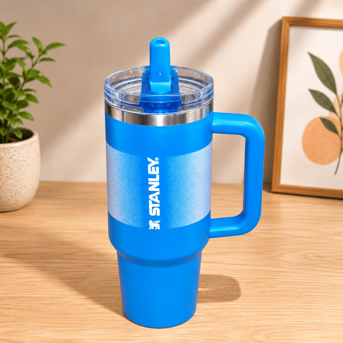40oz Insulated Stainless Steel TumblerTumblersCrafty CuddleA bright blue Stanley tumbler with a matching blue lid and handle, standing on a light wood tabletop. The background shows a softly blurred indoor setting with a small plant and framed artwork.Blue1.18L tumblertumbler with straw