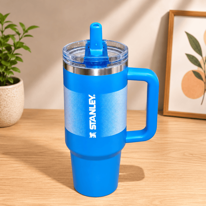 40oz Insulated Stainless Steel TumblerTumblersCrafty CuddleA bright blue Stanley tumbler with a matching blue lid and handle, standing on a light wood tabletop. The background shows a softly blurred indoor setting with a small plant and framed artwork.Blue1.18L tumblertumbler with straw