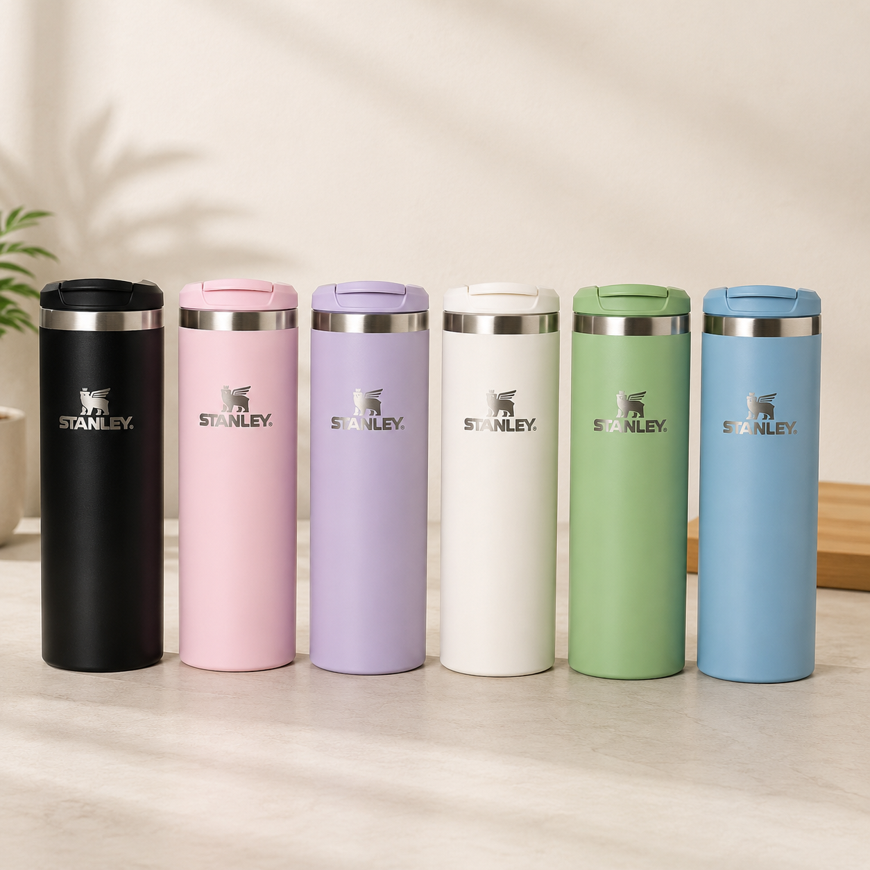 Six Stanley travel tumbler thermoses in different colors (black, pink, lavender, off-white, sage green, and sky blue) lined up on a light wooden surface against a warm beige background.