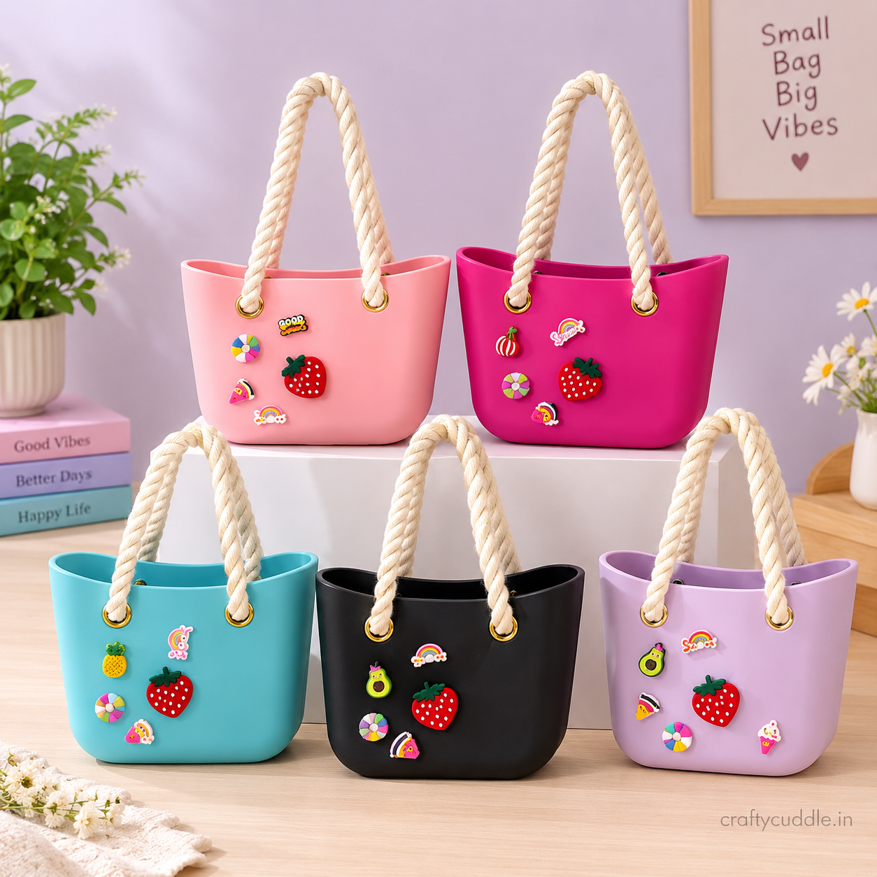 Five small tote bags in pink, magenta, turquoise, black, and lavender, each decorated with colorful fruit charms and featuring thick cream rope handles, arranged on a tabletop with a lavender wall background.