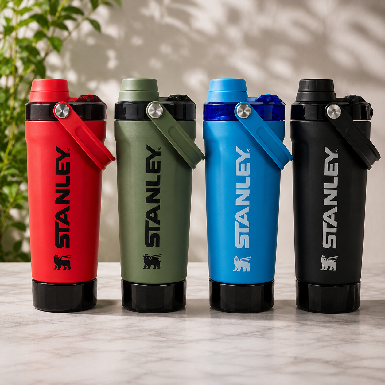 Four Stanley water bottles in red, olive green, blue, and black, lined up on a light marble surface with a soft shadow and blurred greenery in the background.