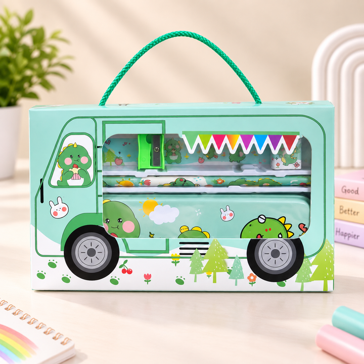 A mint green cartoon stationery set in a vehicle-shaped box with a green rope handle. The box features a cute green dinosaur character and colorful decorative details, set against a softly blurred desk background with a plant and books.