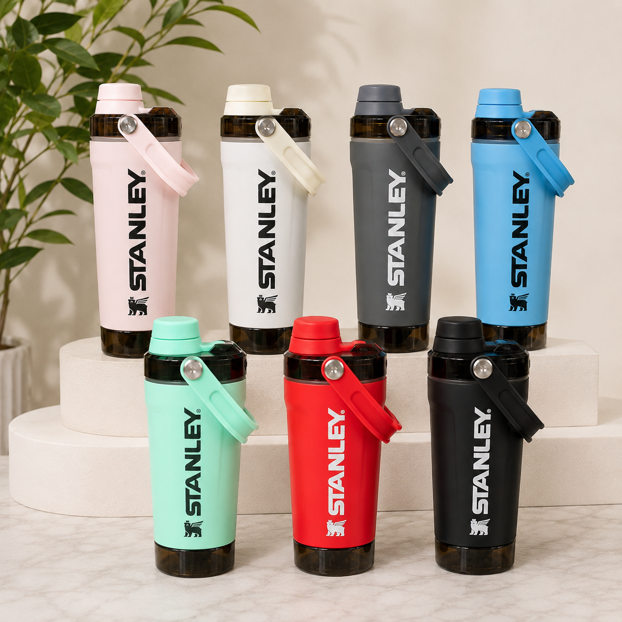 Six Stanley shaker bottles in different colors (pink, white, dark gray, light blue, mint green, red, and black) arranged on white risers and a marble surface with a green plant in the background.
