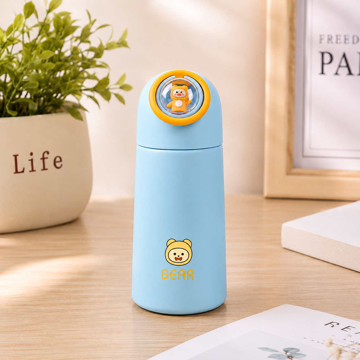 A light blue glass water bottle with a yellow bear-themed lid and a yellow bear logo on the front, standing on a wooden tabletop in a styled indoor setting with a potted plant and framed art in the background.