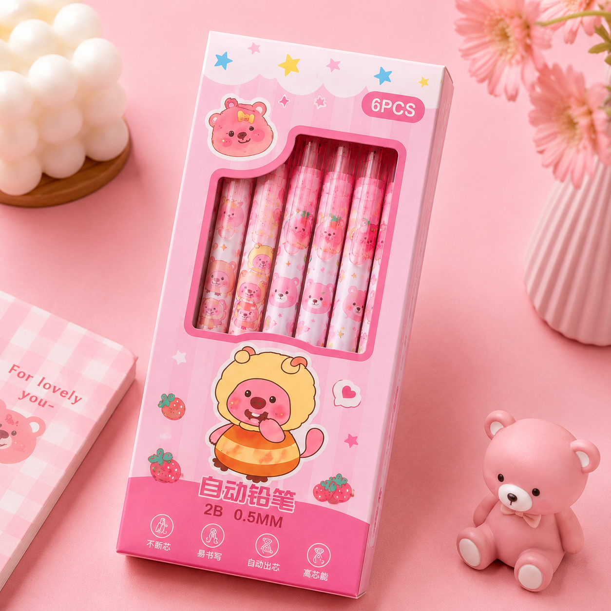 A pink cartoon-themed 6-pack box of mechanical pencils, displayed on a pink background with decorative props including flowers, a candle, and a small teddy bear.