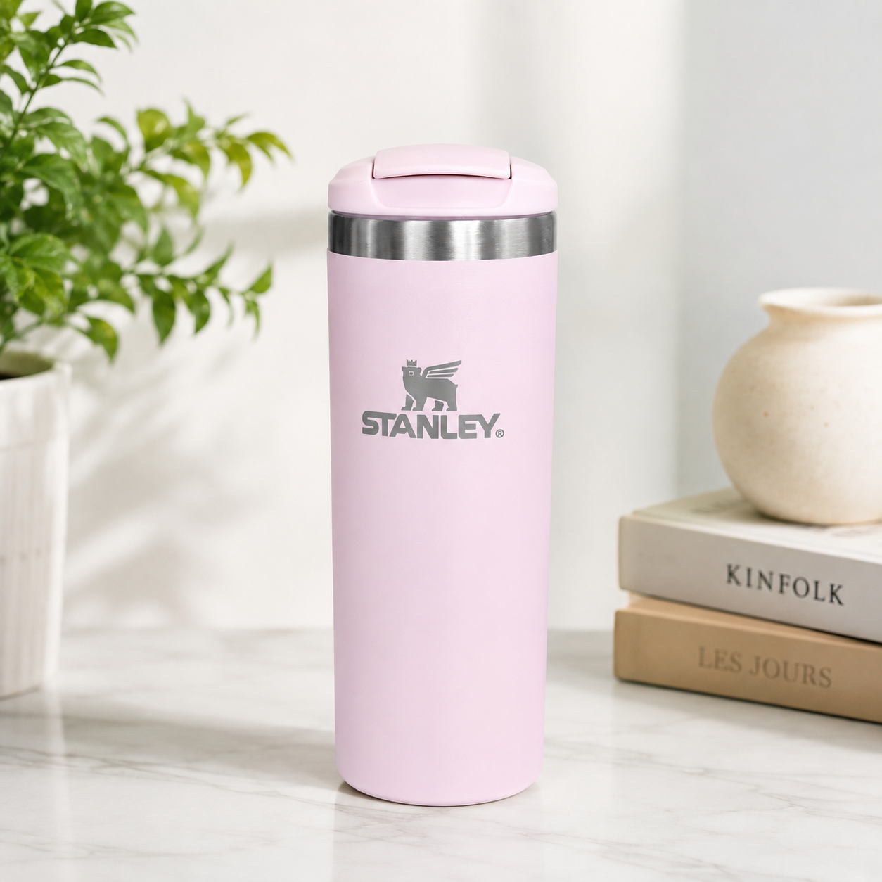 A light pink Stanley travel tumbler with a stainless steel rim, standing on a marble surface next to a potted plant and a stack of books.