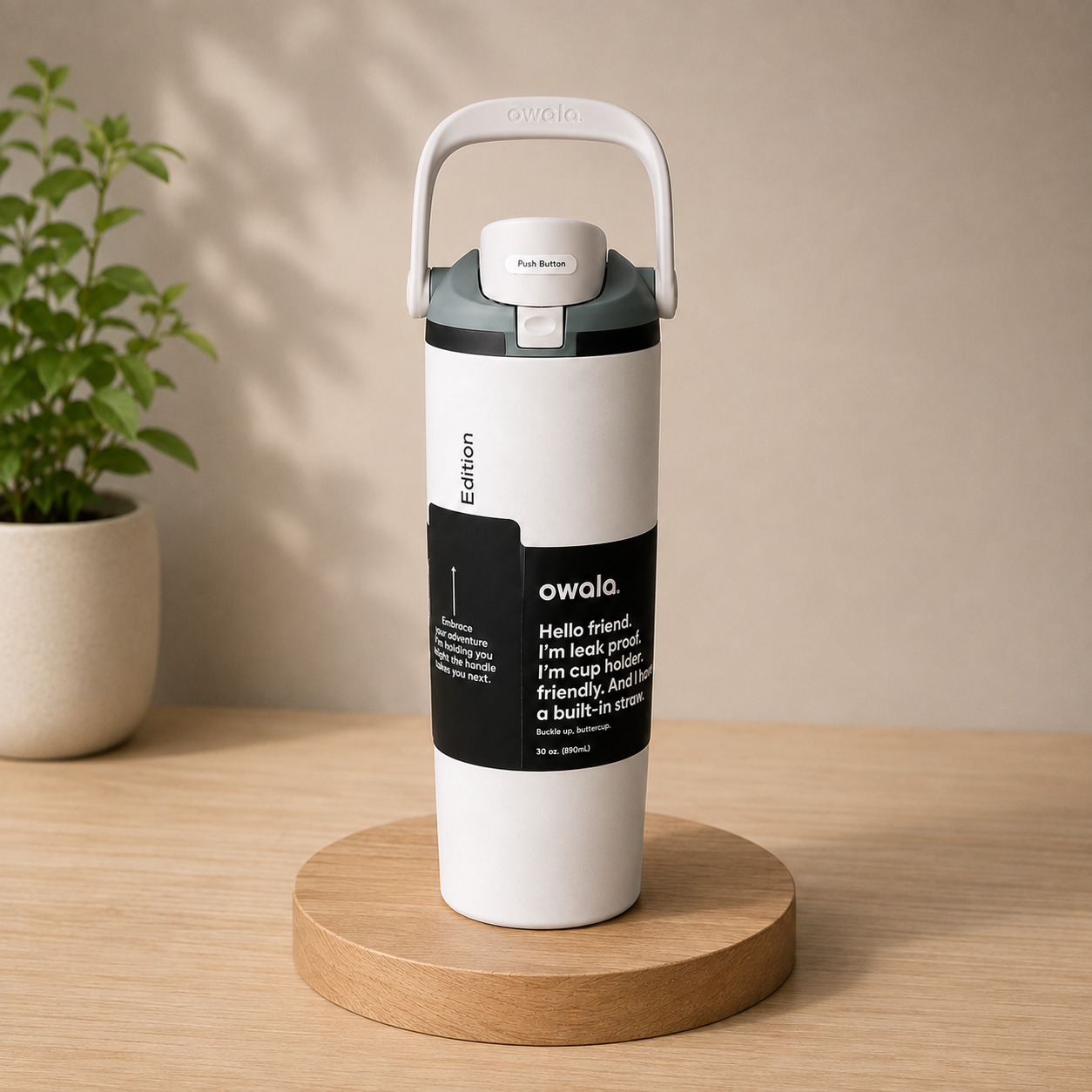 A white Owala insulated tumbler with a gray lid and carry loop, standing on a round wooden coaster on a light wood table. A potted plant is visible in the background against a beige wall.