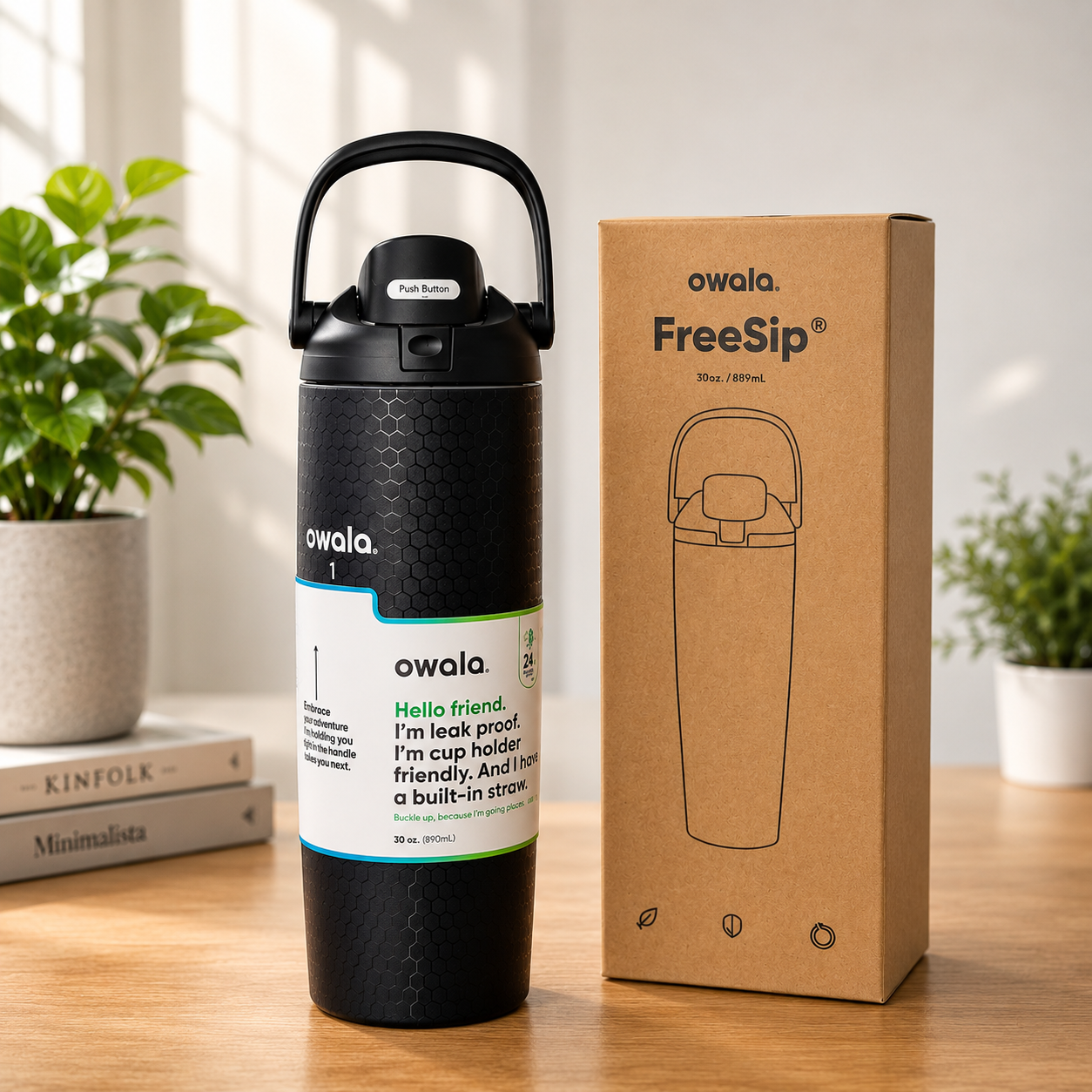 A black Owala FreeSip insulated tumbler with a carry loop and a white label stands beside its brown cardboard packaging box on a wooden table, with potted plants and books in the background.