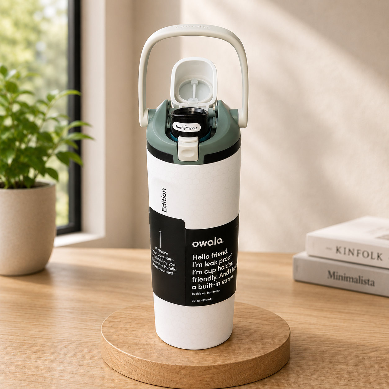 A white Owala water bottle with a dark green lid and handle, standing on a round wooden coaster on a wooden table. The background features a soft-focus indoor setting with a potted plant and books.