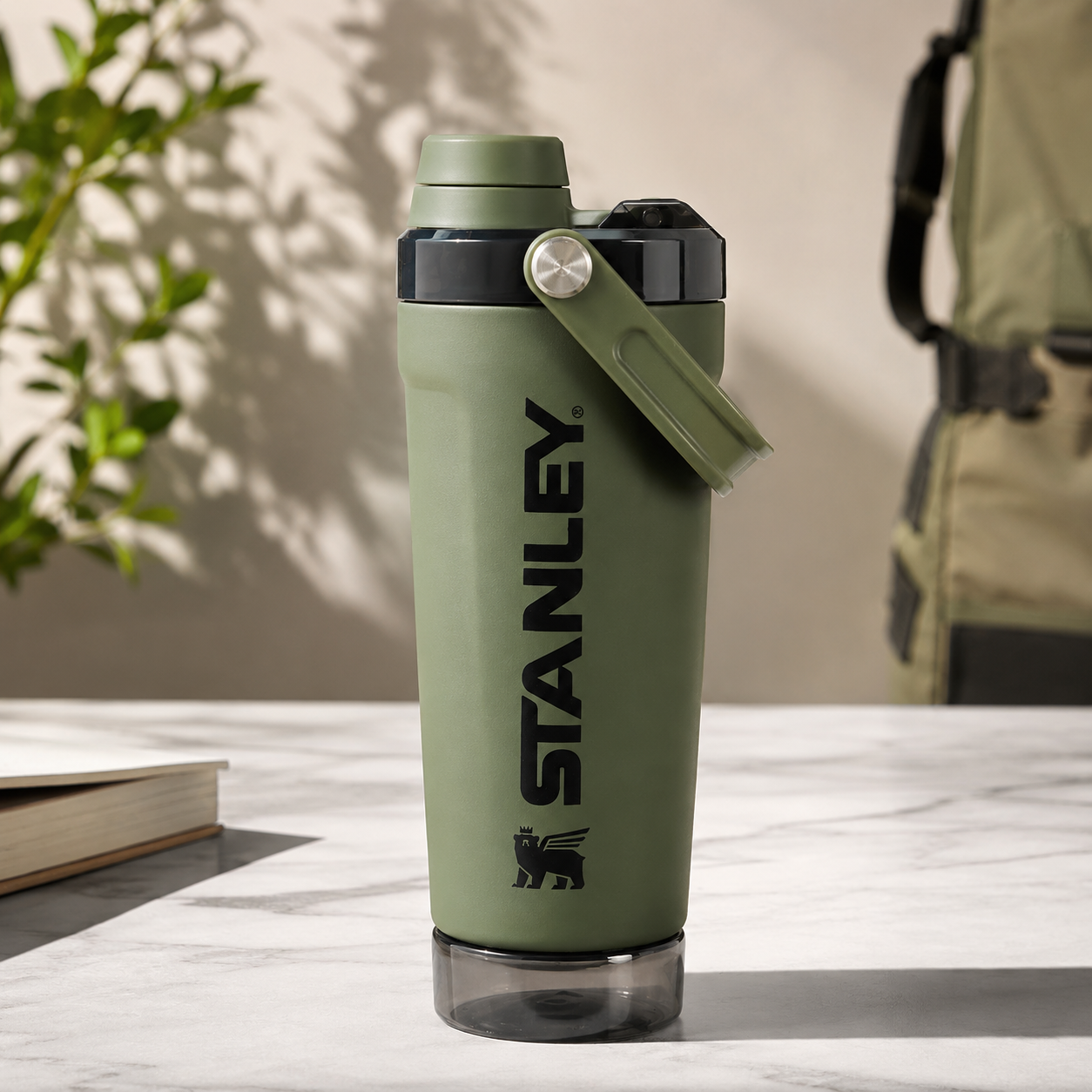 Stanley water bottle in olive green with a black logo and a matching green carrying loop, sitting on a light marble surface with a blurred background.