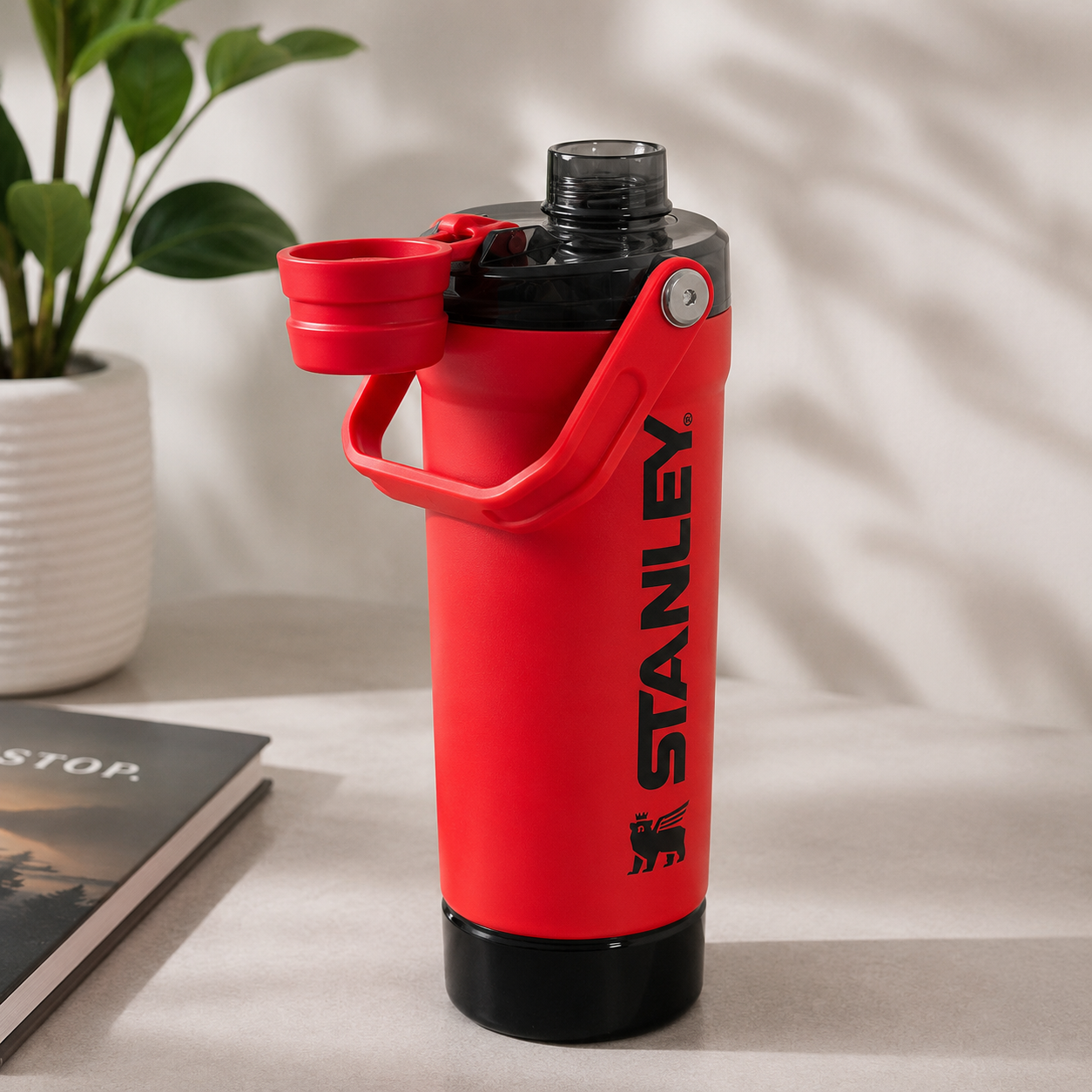 A red Stanley insulated water bottle with a black base and a flip-up lid, shown on a tabletop with a plant and book in the background.
