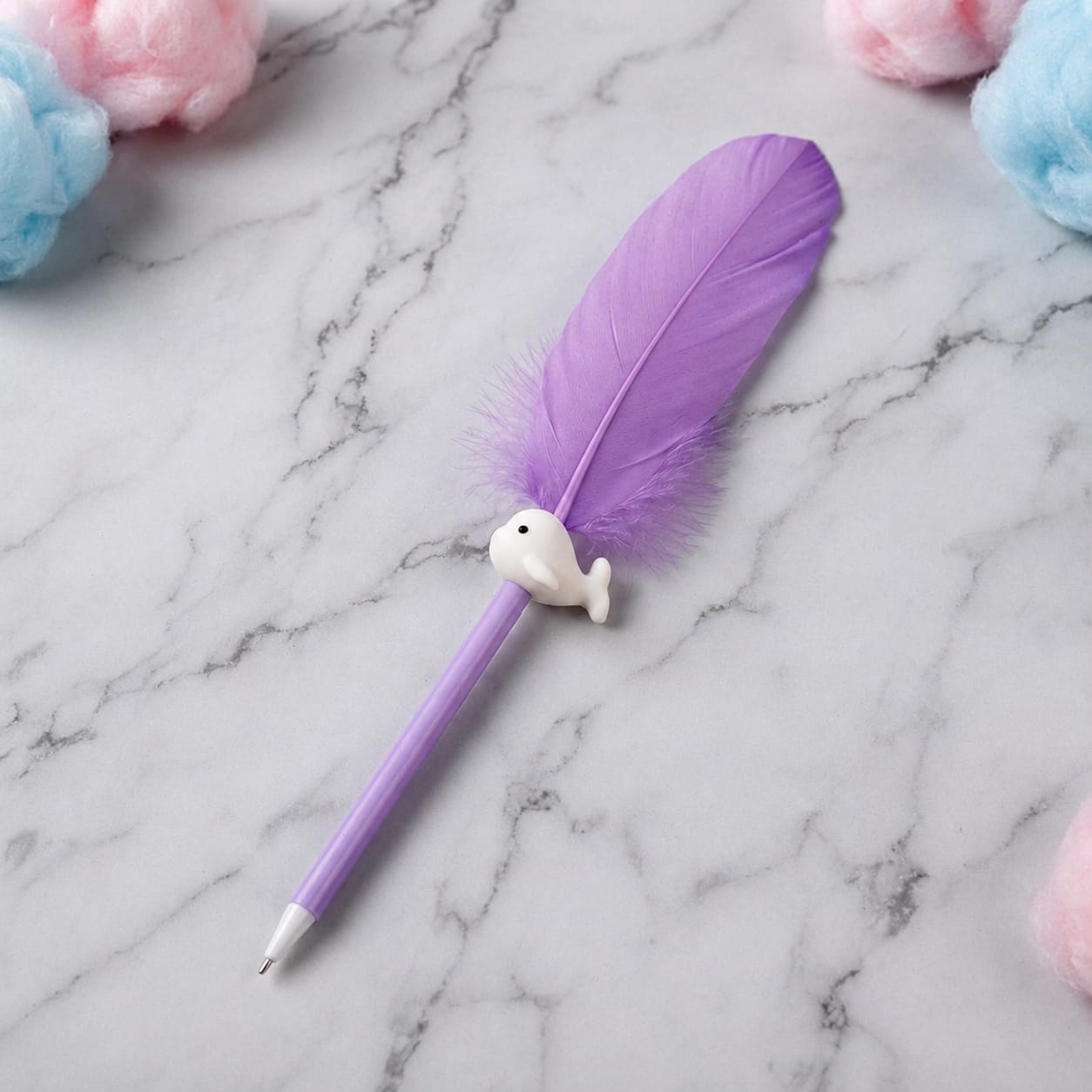 A purple retractable pen with a large purple feather attached near the top and a small white whale-shaped charm, placed on a white marble surface with pastel cotton candy props in the corners.