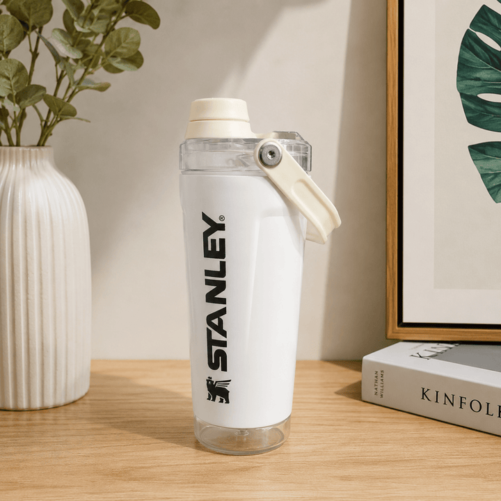 Activate Shaker BottleInsulated Water BottleCrafty CuddleA white Stanley water bottle with a clear lid and cream-colored carrying loop, standing on a wooden table next to a white vase with green leaves and a framed print.WhiteShakerBottle ProteinShaker GymBottle FitnessEssentials WaterBottle WorkoutGear LeakproofBottle BPAFree SportsBottle Hydration