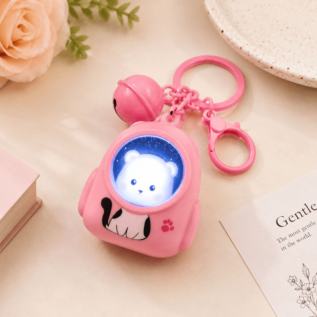 Cute Cartoon LED Keychain with BellKeychainsCrafty CuddleA pink astronaut-shaped keychain toy with a glowing blue window showing a white bear face, attached to a pink bell and key rings, styled on a light beige surface with decorative flowers and a book.CuteKeychain CartoonKeychain BagCharm KeychainCollection KawaiiAccessories CuteAccessories GiftIdeas ReturnGifts StationeryLovers CraftyCuddleStationeryAddict