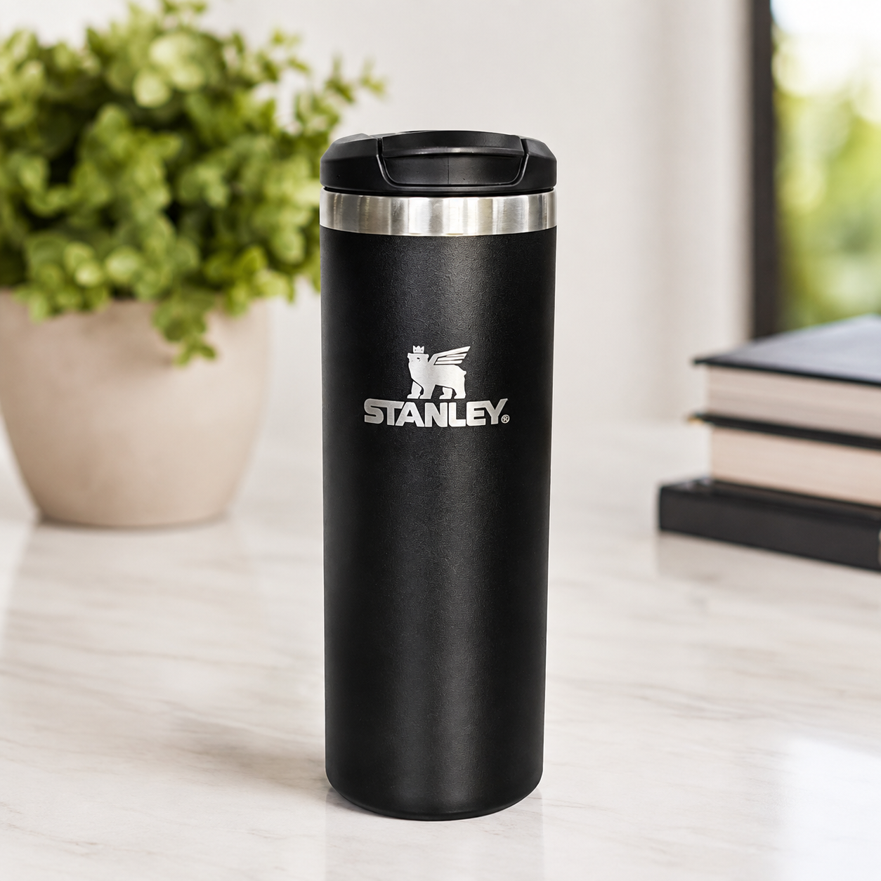 A black Stanley travel tumbler with a stainless steel rim and a black lid, standing on a light marble countertop with a blurred potted plant and books in the background.