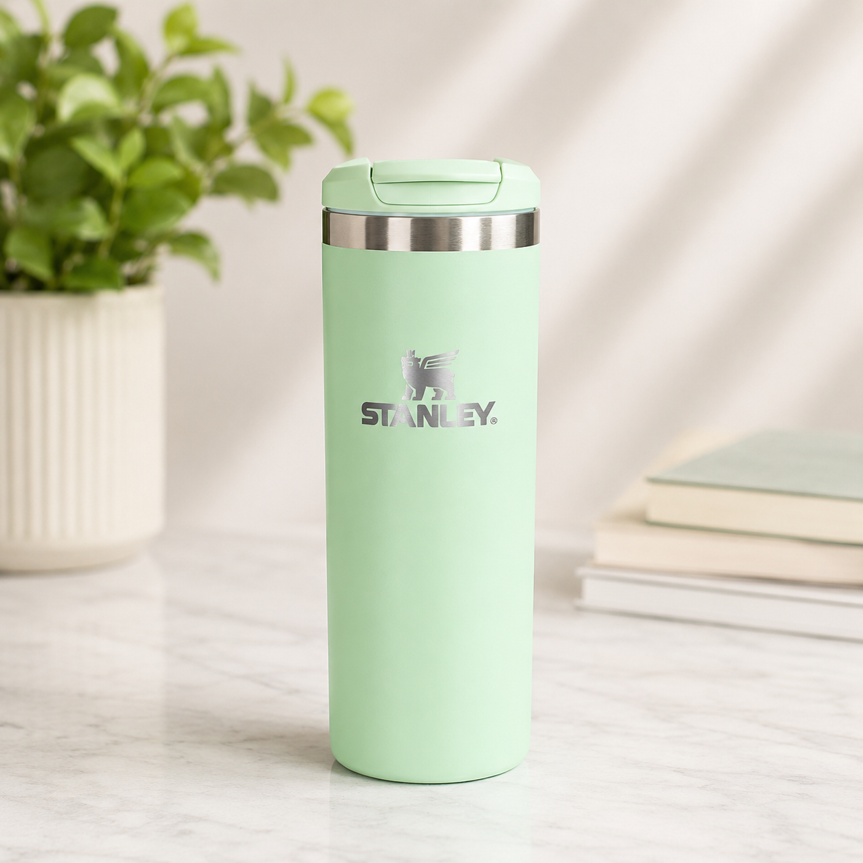A mint green Stanley travel tumbler with a stainless steel lid, standing on a white marble surface with a potted plant and books in the background.