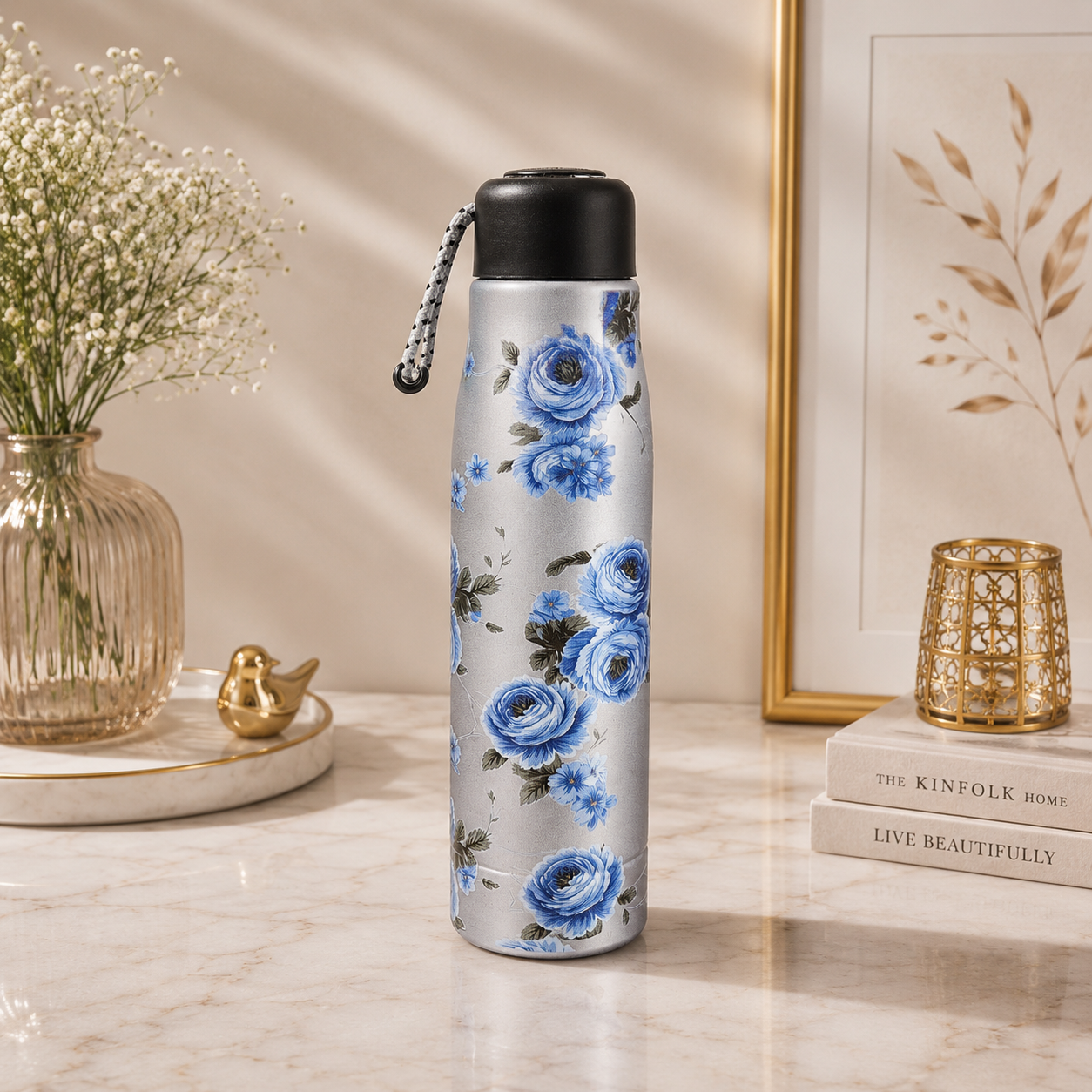 A reusable insulated bottle with a blue floral print on a silver background, featuring a black lid and a silver chain strap, displayed on a marble tabletop with decorative props.