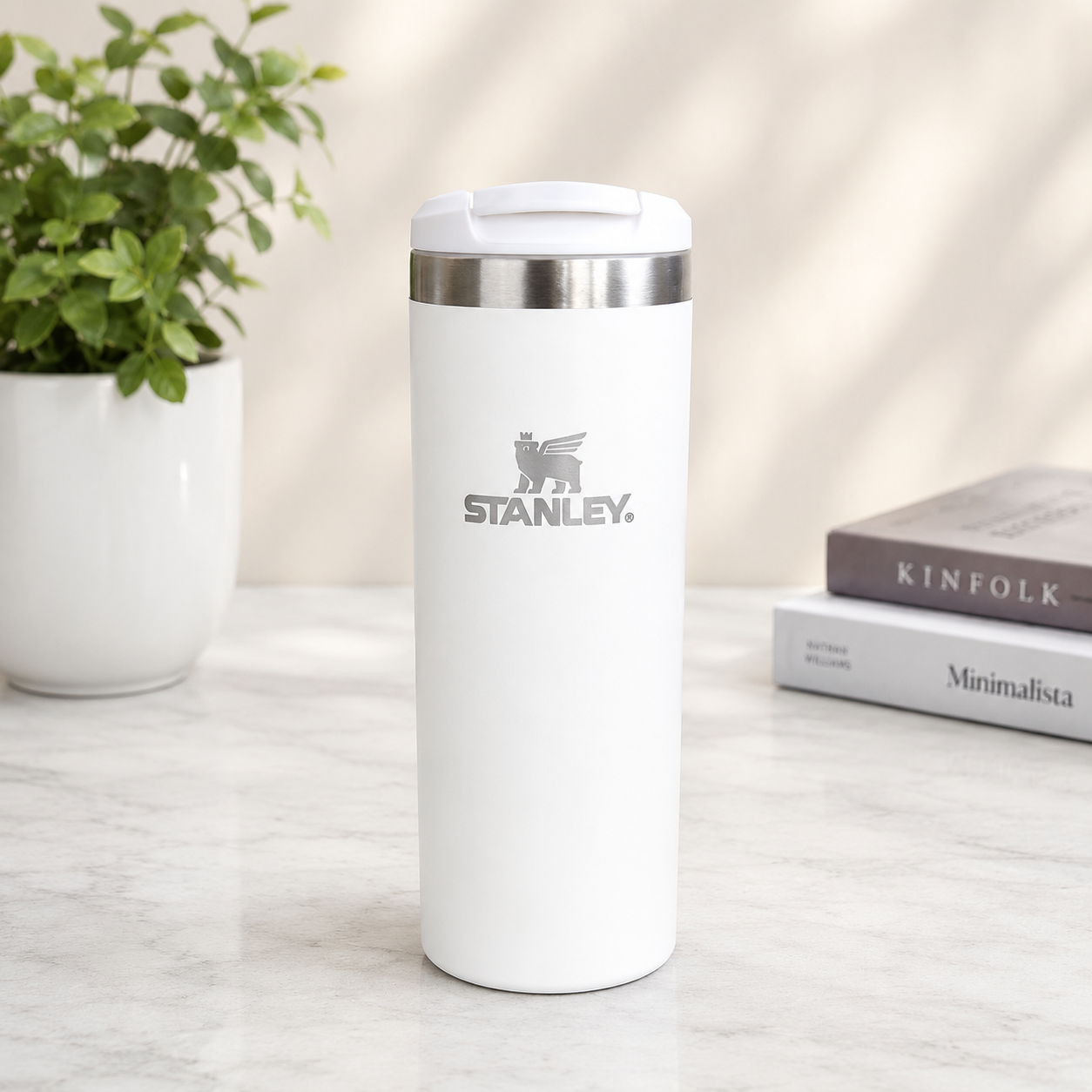 A white Stanley travel tumbler with a stainless steel rim and a gray logo on the front, standing on a light marble surface with a potted plant and books in the background.