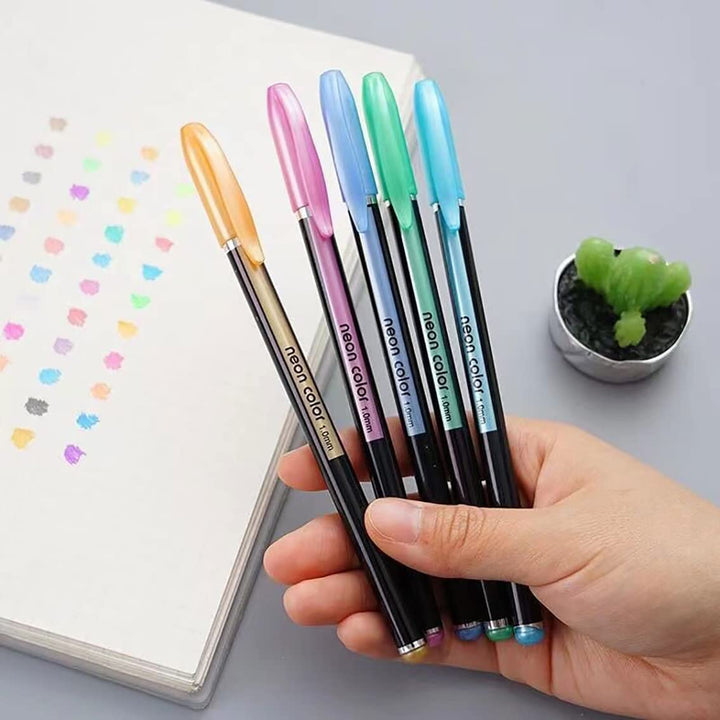 Hand holding five colorful pens next to a notebook with a small plant in the background
