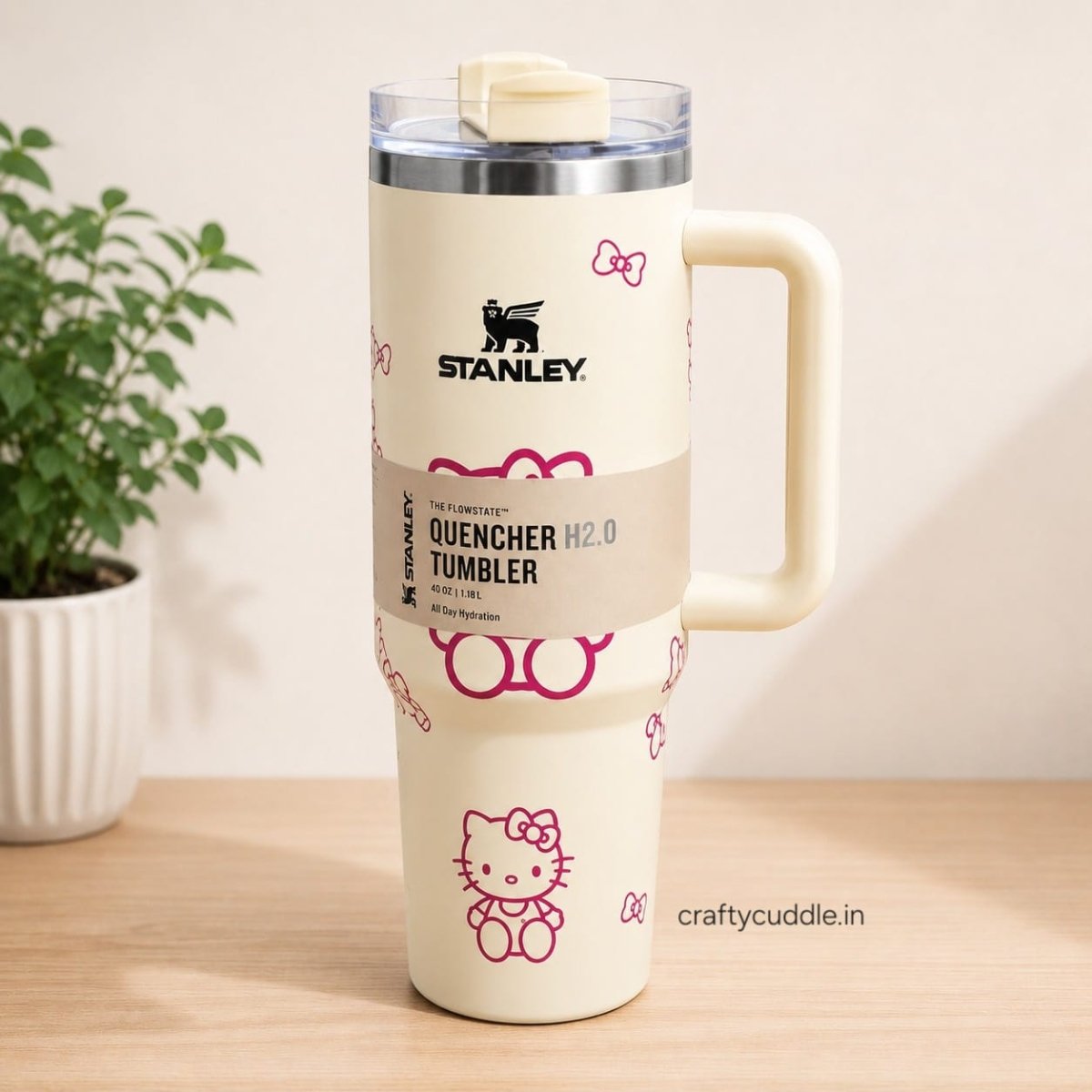 Hello Kitty Stainless Steel TumblerTumblersCrafty CuddleA cream-colored Stanley Quencher H2.0 travel tumbler with a Hello Kitty design, featuring a handle, clear lid, and a paper band label, set on a wooden table next to a potted plant.CreamHello Kitty Tumbler