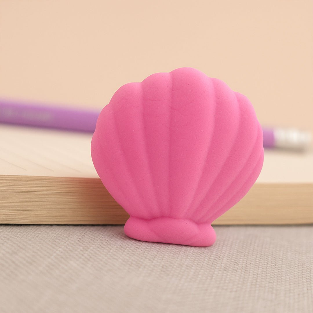 Pink shell-shaped eraser on a beige surface with a pen in the background