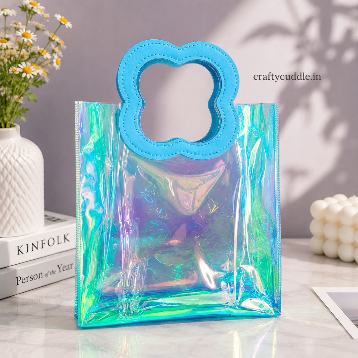 Pretty Shimmer Tote BagTransparent bagCrafty CuddleAn iridescent holographic clear tote bag with a bright blue cloud-shaped handle, displayed on a marble surface with decorative props including a vase of daisies, books, and a candle.BlueGiftHampers GiftPackaging GiftBag IridescentBag GiftIdeas ReturnGifts PartyFavours BirthdayGifts FestiveGifting GiftWrap TrendyBags ReusableBag