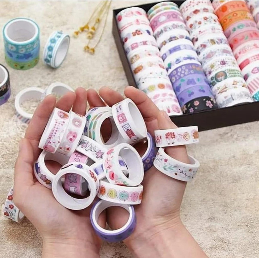 Hand holding colorful washi tape rolls with a box of additional tape in the background.